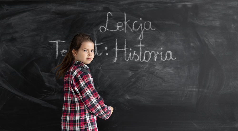 Dziewczynka przy szkolnej tablicy, zapisująca temat lekcji Historia